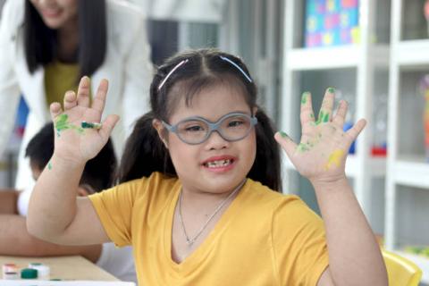 Image of student with disabilities holding up her hands with paint on them. She is wearing a yellow shirt, glasses, and is smiling. 