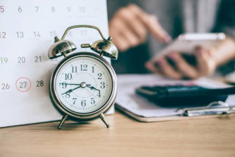 Image of alarm clock placed in front of a calendar. Behind that is an image of a person's hands; the person is using their phone or other device. 