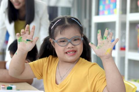 Image of student with disabilities holding up her hands with paint on them. She is wearing a yellow shirt, glasses, and is smiling.